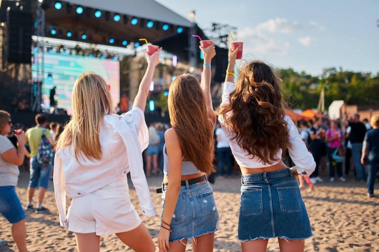 Three girls with their backs facing the camera holding up their cups as they cheer at a music festival on the beach.
