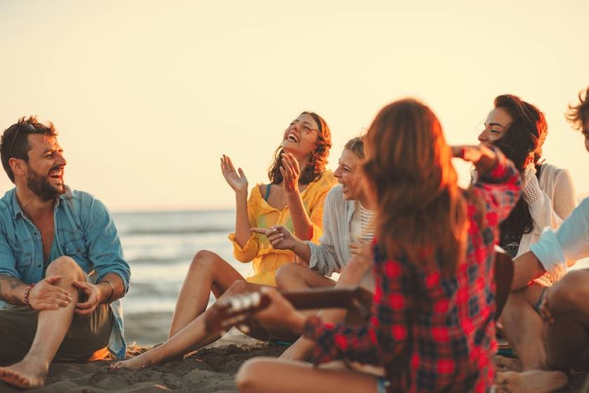 Happy friends sitting on the beach singing and playing guitar during the sunset.